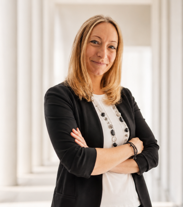 Andrea Höfs mit blonden Haaren, schwarzem Blazer und Perlenkette steht selbstbewusst mit verschränkten Armen in einem architektonischen Gang mit weißen Säulen.