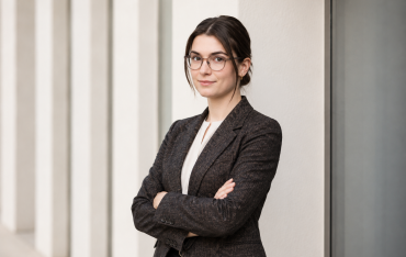 Frau mit Brille und verschränkten Armen in einem dunklen Blazer vor einer modernen Gebäudefassade