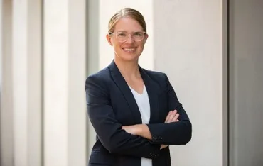 Jennifer in einem dunklen Blazer und mit Brille steht mit vor der Brust verschränkten Armen vor einer hellen Wand.