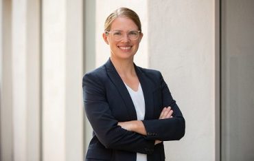 Porträt einer Expertin in Business-Kleidung mit Brille und verschränkten Armen vor hellem Hintergrund.
