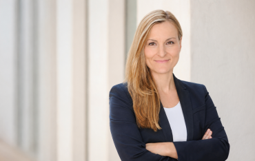 Porträt von Carola Wolf mit blondem, langem Haar und dunklem Blazer vor heller Kulisse.