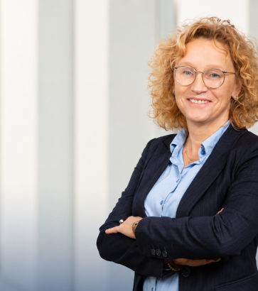 Porträt der Trainerin Maike Grünebaum mit Locken, Brille und blauem Hemd im Business-Look.