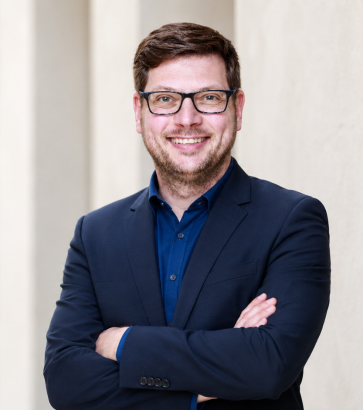 Porträt des Trainers Richard Röhlig mit Brille, blauem Hemd und dunklem Sakko vor einer hellen Fassade.