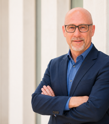 Portretul antrenorului Thomas Zydeck cu ochelari, barbă și cămașă albastră în fața unei arhitecturi moderne cu coloane.