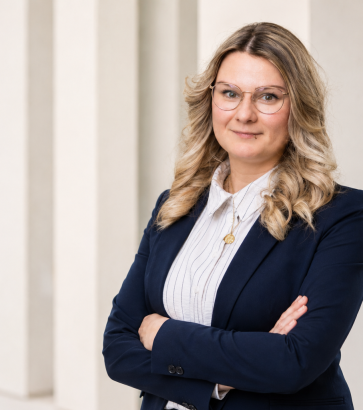 Porträt von Aniko Koch mit Brille, heller Bluse und dunklem Blazer vor heller Wand.