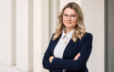 Porträt von Aniko Koch mit Brille, heller Bluse und dunklem Blazer vor heller Wand.