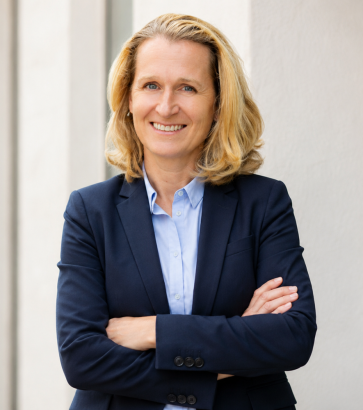 Porträt der Trainerin Karen Everding mit blauem Hemd und dunklem Blazer vor einer Säulenwand.
