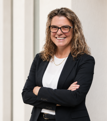 Porträt der Trainerin Simone Hoffmann mit Brille und schwarzem Blazer vor einer hellen Säulenwand.